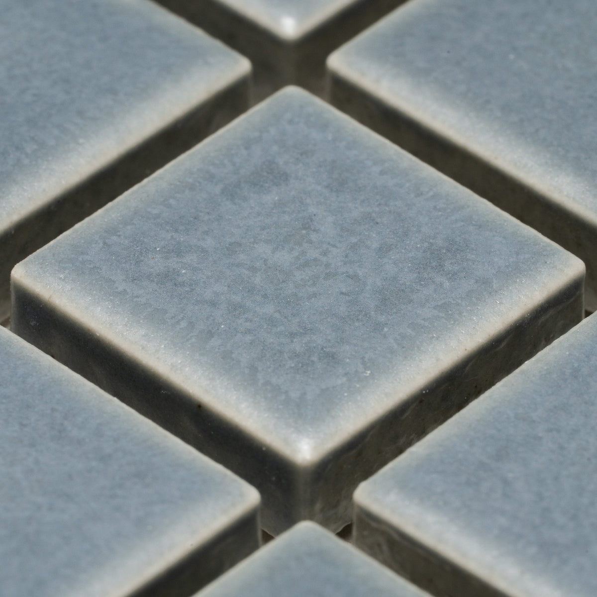 Close-up of Loch 1x1 gray ceramic tiles by Zia Tile, featuring a slightly textured matte surface, square shape, and visible raised edges with gaps arranged in a grid pattern.