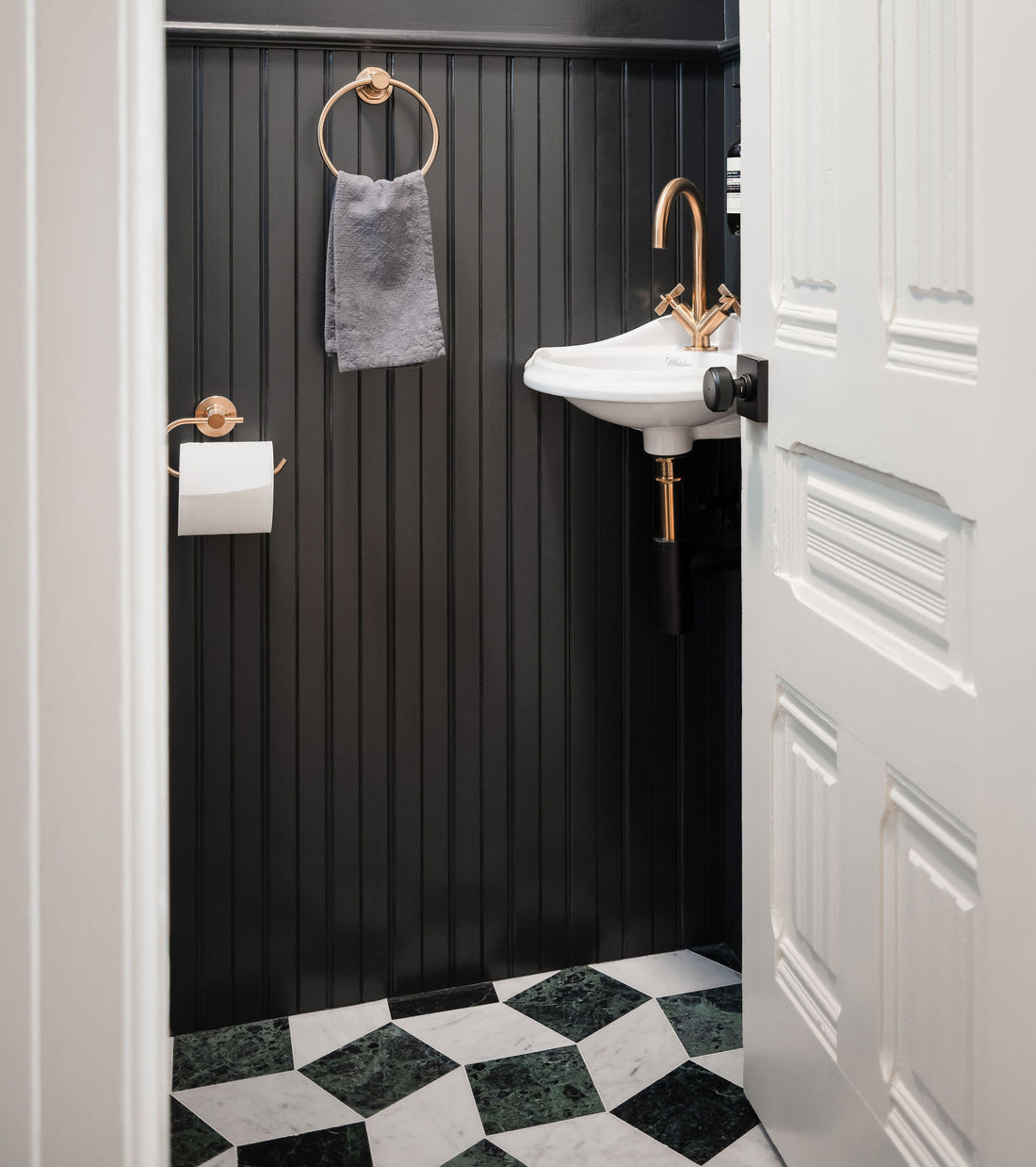 A small bathroom with black beadboard walls, a white wall-mounted sink with gold hardware, and a checkered Zia Tile Lombardy in Carrara + Verde Alpi marble floor in green and white.