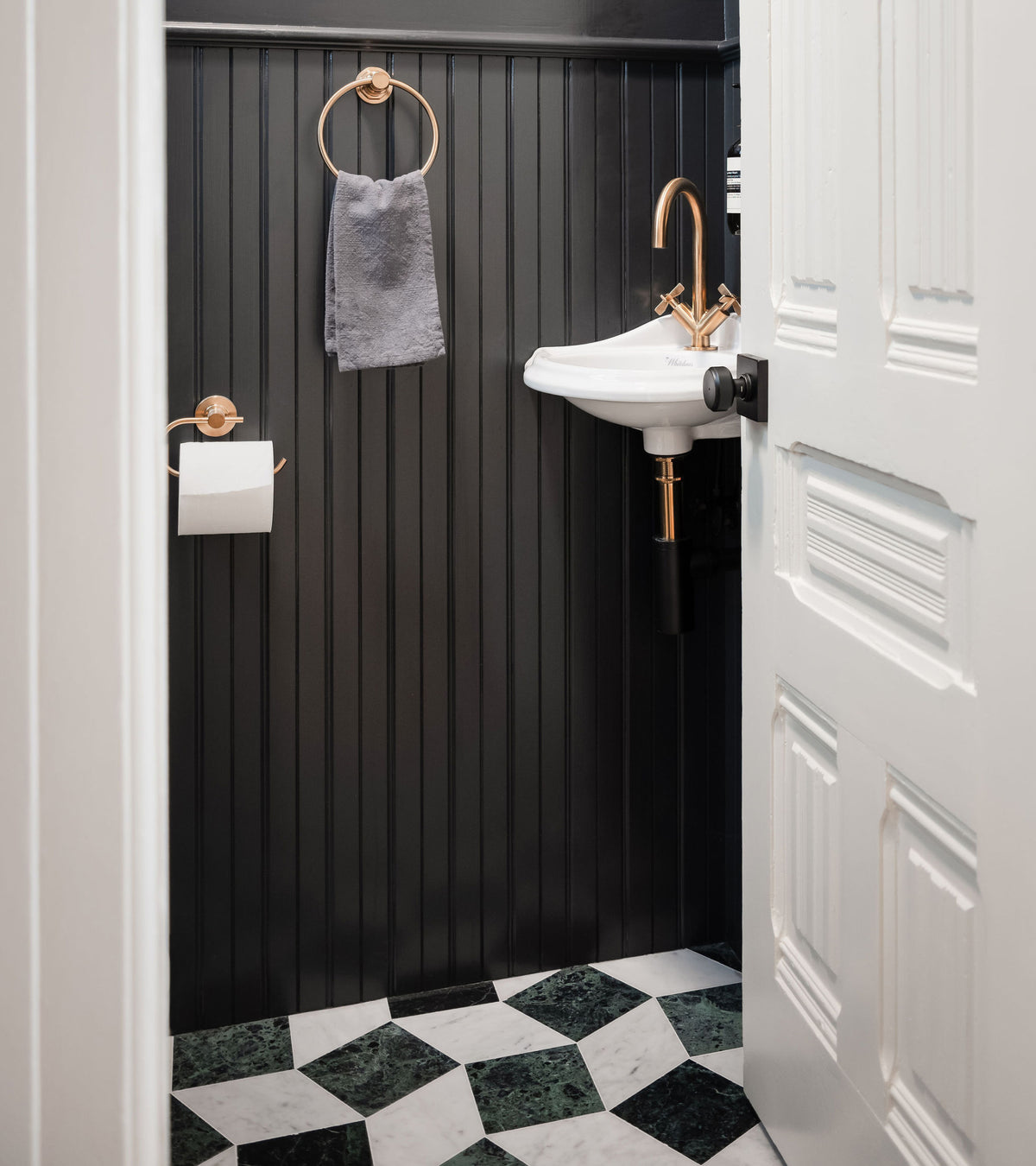 A small bathroom with black beadboard walls, a white wall-mounted sink with gold hardware, and a checkered Zia Tile Lombardy in Carrara + Verde Alpi marble floor in green and white.
