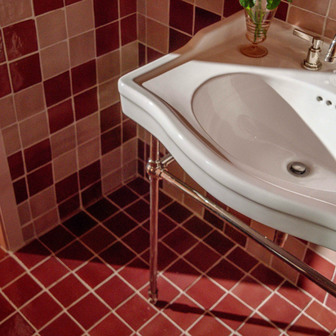 A curved white sink with metal legs is mounted in a bathroom corner featuring Zia Tile’s Lupe 4x4 red and pink handmade cotto and Mexican ceramic tiles. A green potted plant sits on the countertop beside the faucet.