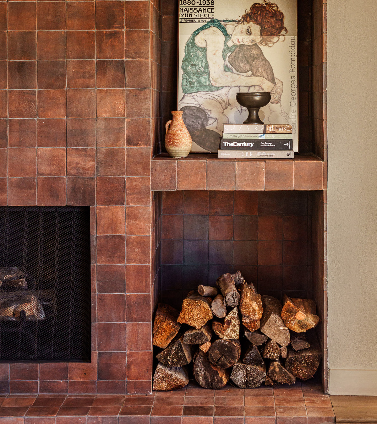 A brown handmade 4x4 Square + Madera Cotto tiled fireplace by Zia Tile features a niche of stacked firewood and a shelf above with framed art, vases, and books, creating a warm, cozy, and artistic atmosphere.