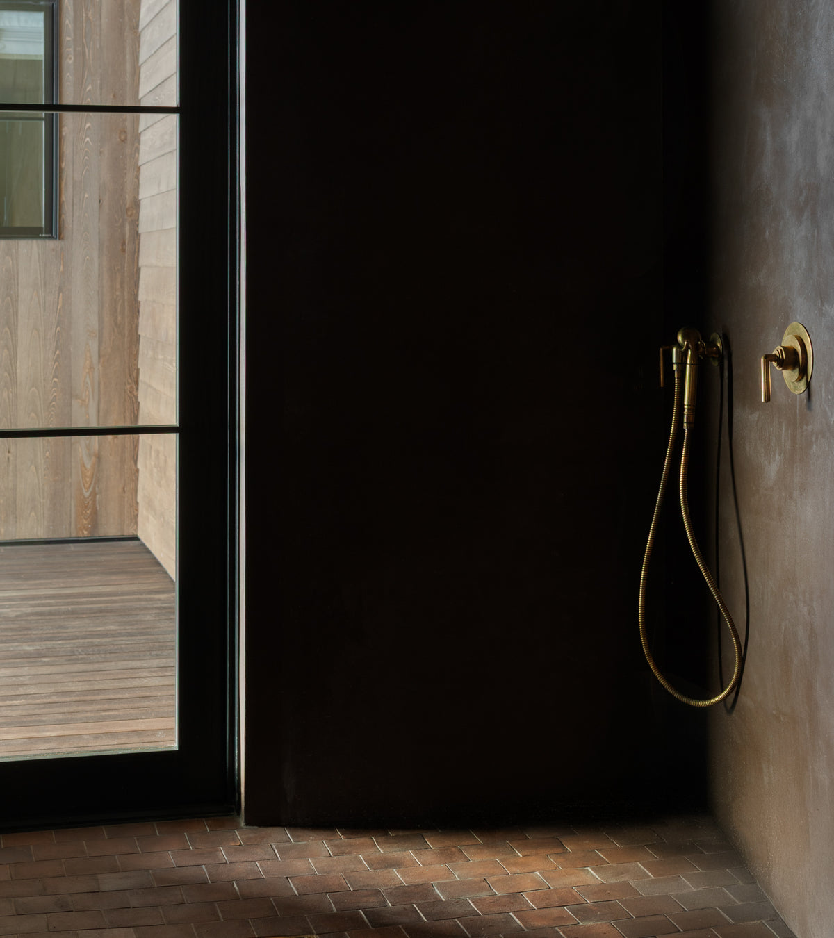 A minimalist shower with a brass handheld showerhead stands by a glass door, opening to a wooden deck with vertical wood siding and Zia Tile’s 4x4 Square + Madera handmade tiles inspired by San Miguel de Allende.