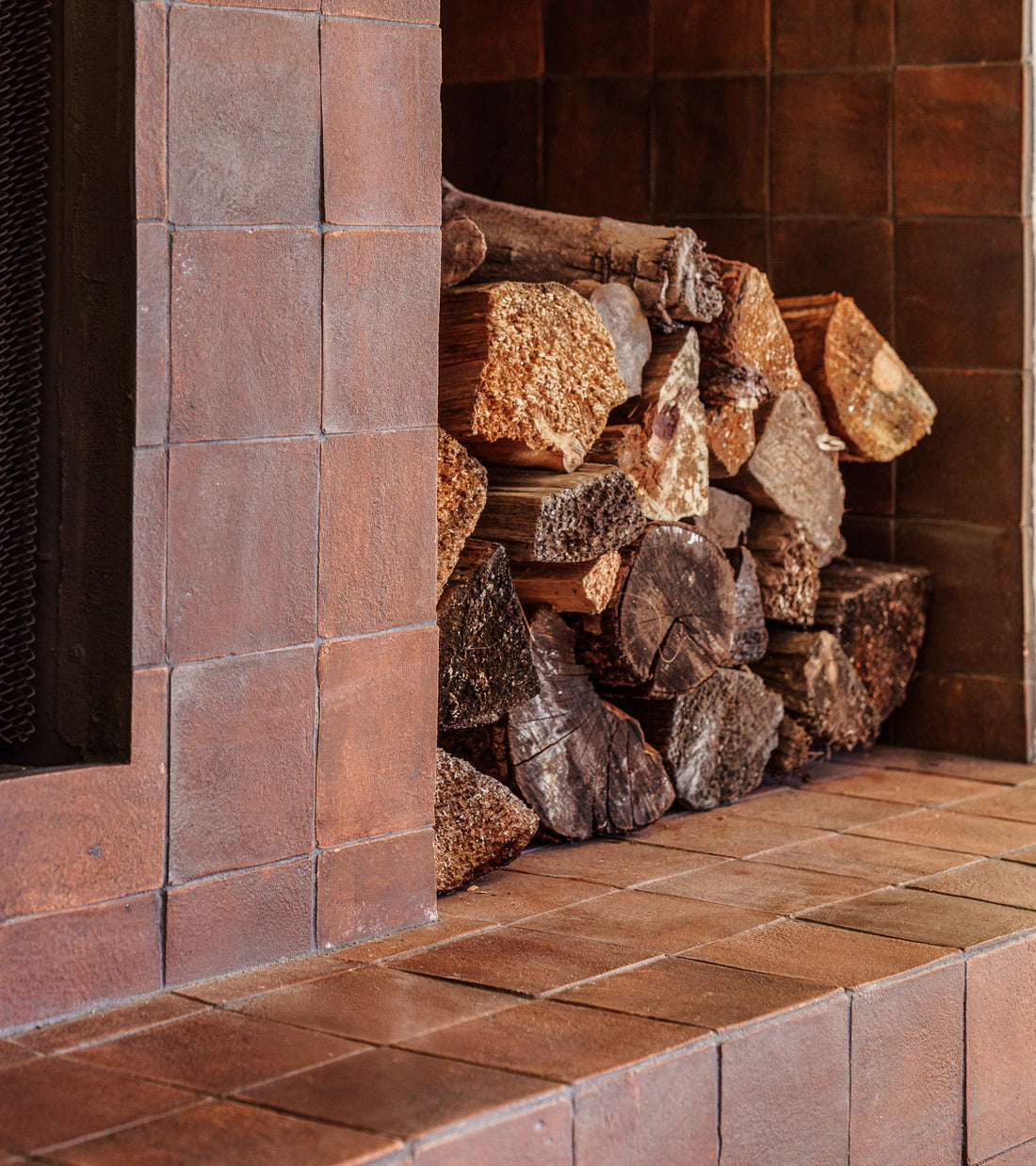 A stack of firewood sits inside a fireplace, framed by warm, reddish-brown 4x4 Square + Madera tiles from Zia Tile, reflecting the timeless charm and craftsmanship of traditional ceramic design.