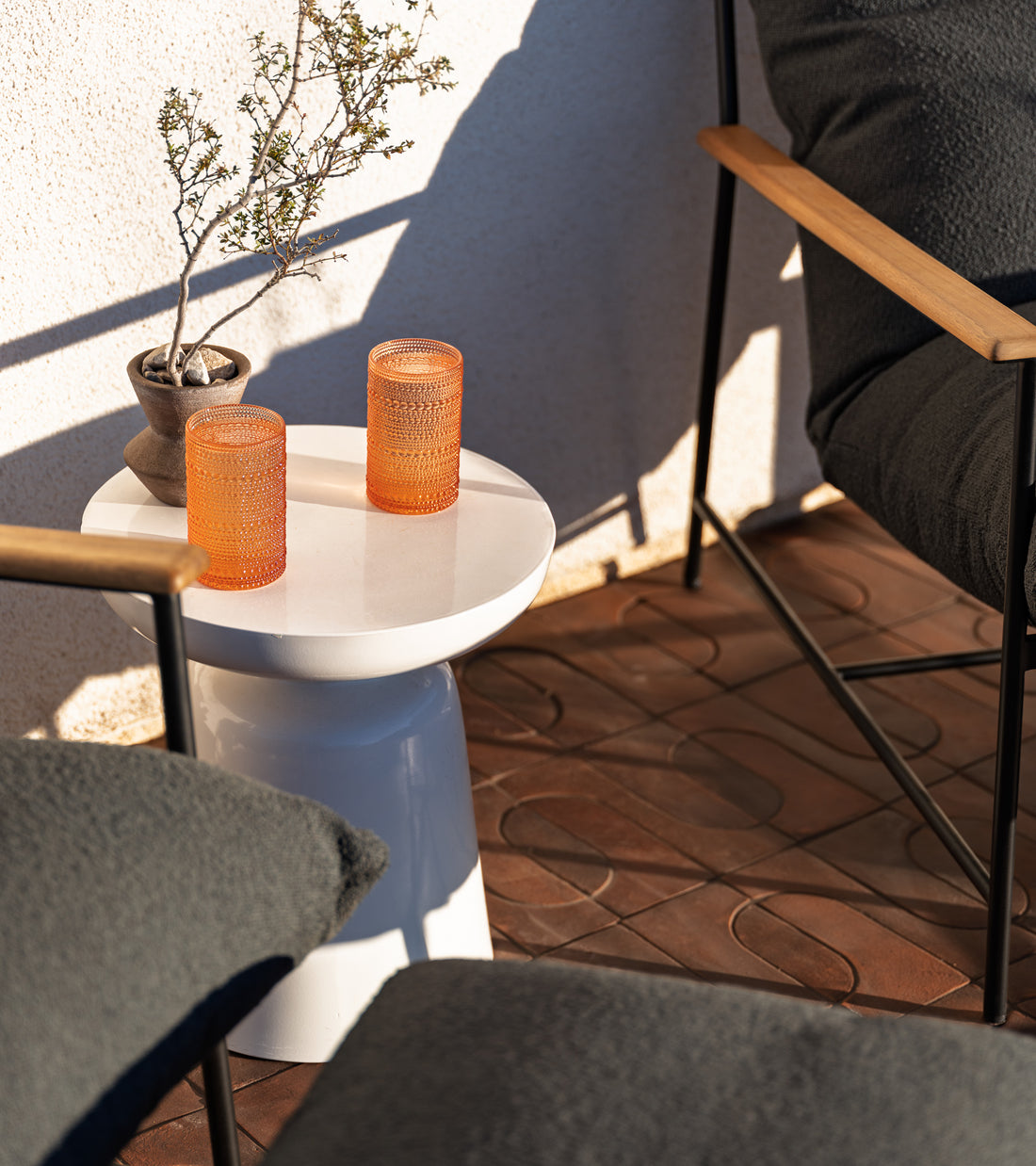 A sunlit patio with Zia Tile’s Durango + Madera patterned brown tiles features a small white side table holding two textured orange glasses and a potted plant, placed between cushioned chairs with wooden armrests.