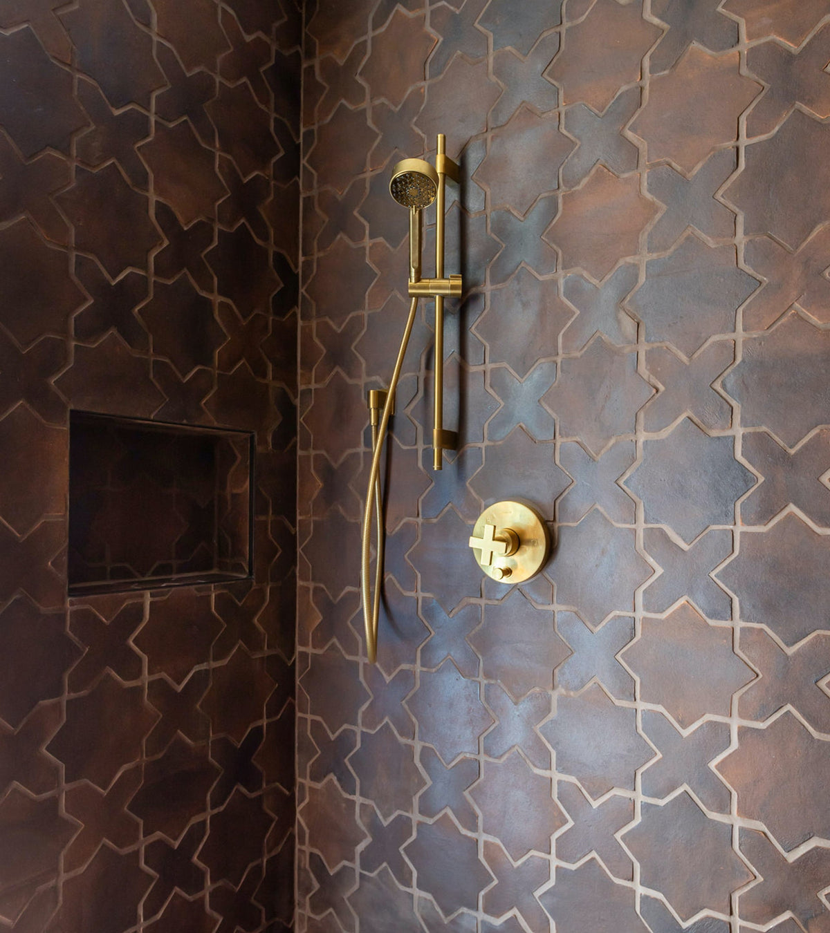 Shower with intricate brown tile walls featuring a geometric pattern, a built-in niche, and gold fixtures including a modern showerhead and control handle.