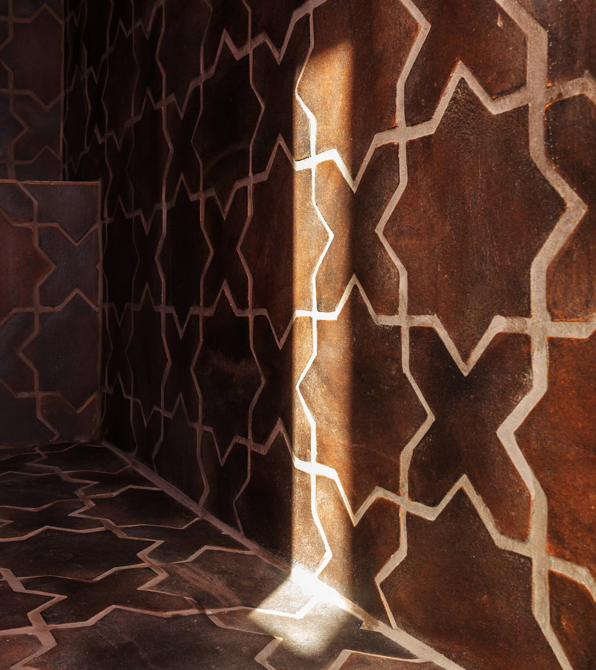 A wall and floor covered with dark tiles featuring a geometric star pattern, illuminated by a narrow beam of sunlight coming through an unseen window.