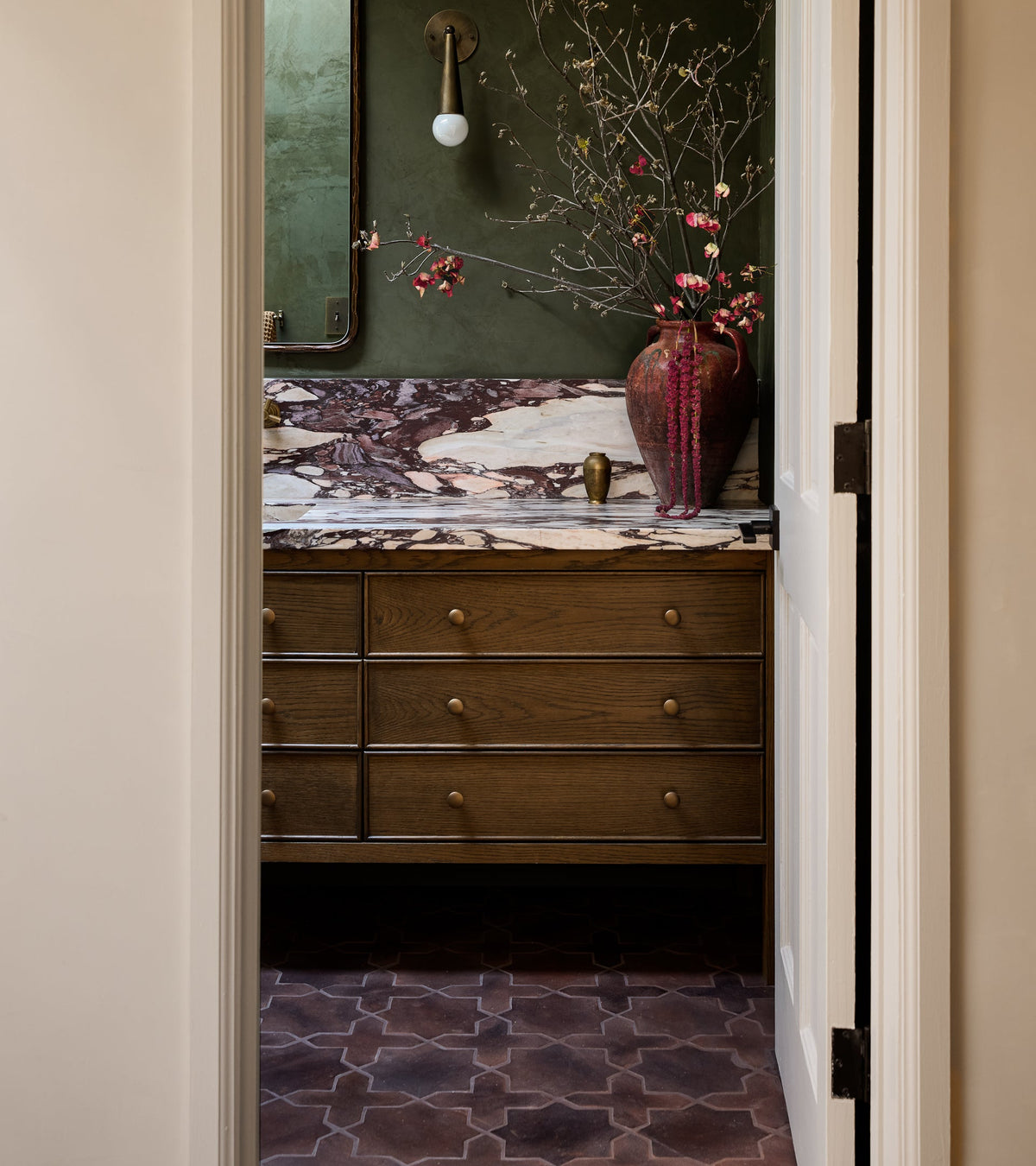 A bathroom with a wooden vanity, marble countertop, brass hardware, and a large red vase with branches and pink flowers. The wall is green with a brass sconce and mirror, seen through a partially open white door.