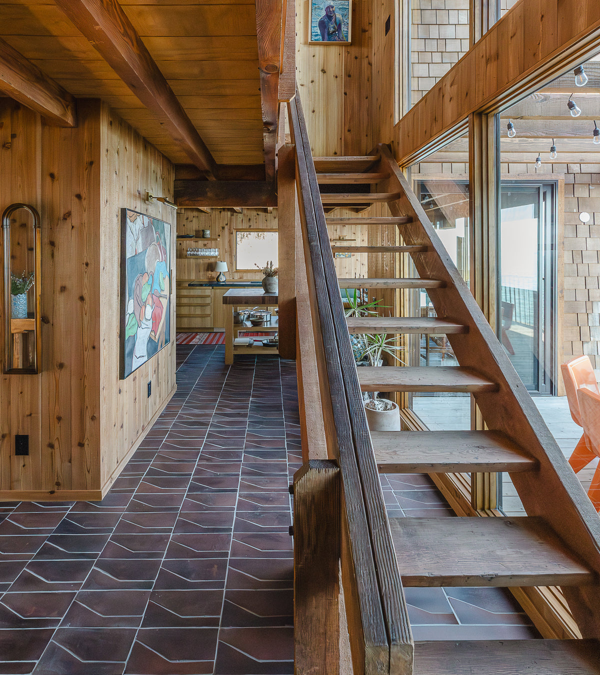 A wooden staircase with open risers leads to an upper level in a cozy, sunlit cabin with wood-paneled walls, large windows, and handmade Zocalo + Madera terra cotta tile flooring by Zia Tile, accented by colorful artwork.