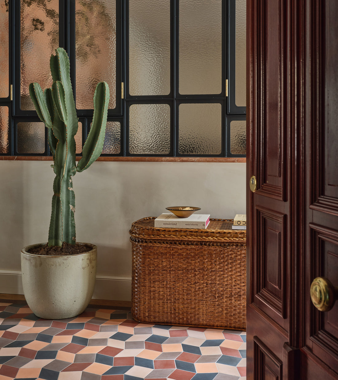 A tall cactus in a white pot sits by a wicker chest under frosted windows; geometric handmade Madrid Set One tile floors from Zia Tile and a partially open wooden door complete the scene.