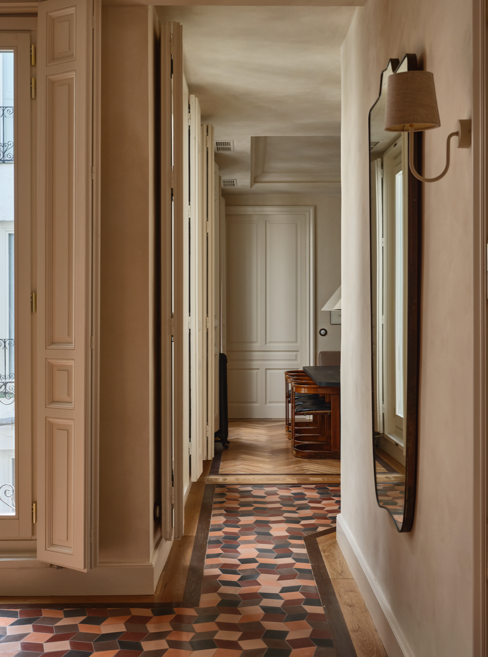 A hallway featuring Zia Tile’s Madrid Set One geometric floor tiles transitions to wood flooring. Tall shuttered windows line the left, with a wall mirror and light fixture on the right, and a wooden cabinet near a white door at the end.