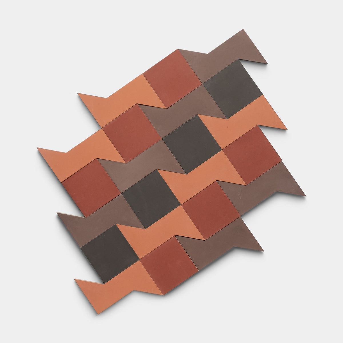 The Manolo Set One, Sample by Zia Tile features interlocking, arrow-shaped tiles in brown, red, and terracotta tones arranged in a staggered geometric pattern on a white background.