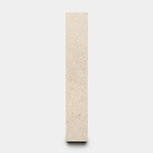 The Mesa 2x12 by Zia Tile is a vertical, rectangular beige stone slab with a smooth surface and subtle natural speckles, shown standing upright against a plain white background.