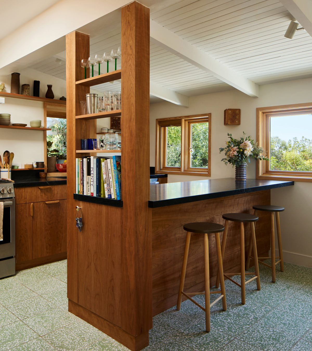 A kitchen with wooden cabinets and an island, featuring a tall open shelf, a black countertop, three wooden stools, and light green speckled Mesquite 12x12 terrazzo tiles.