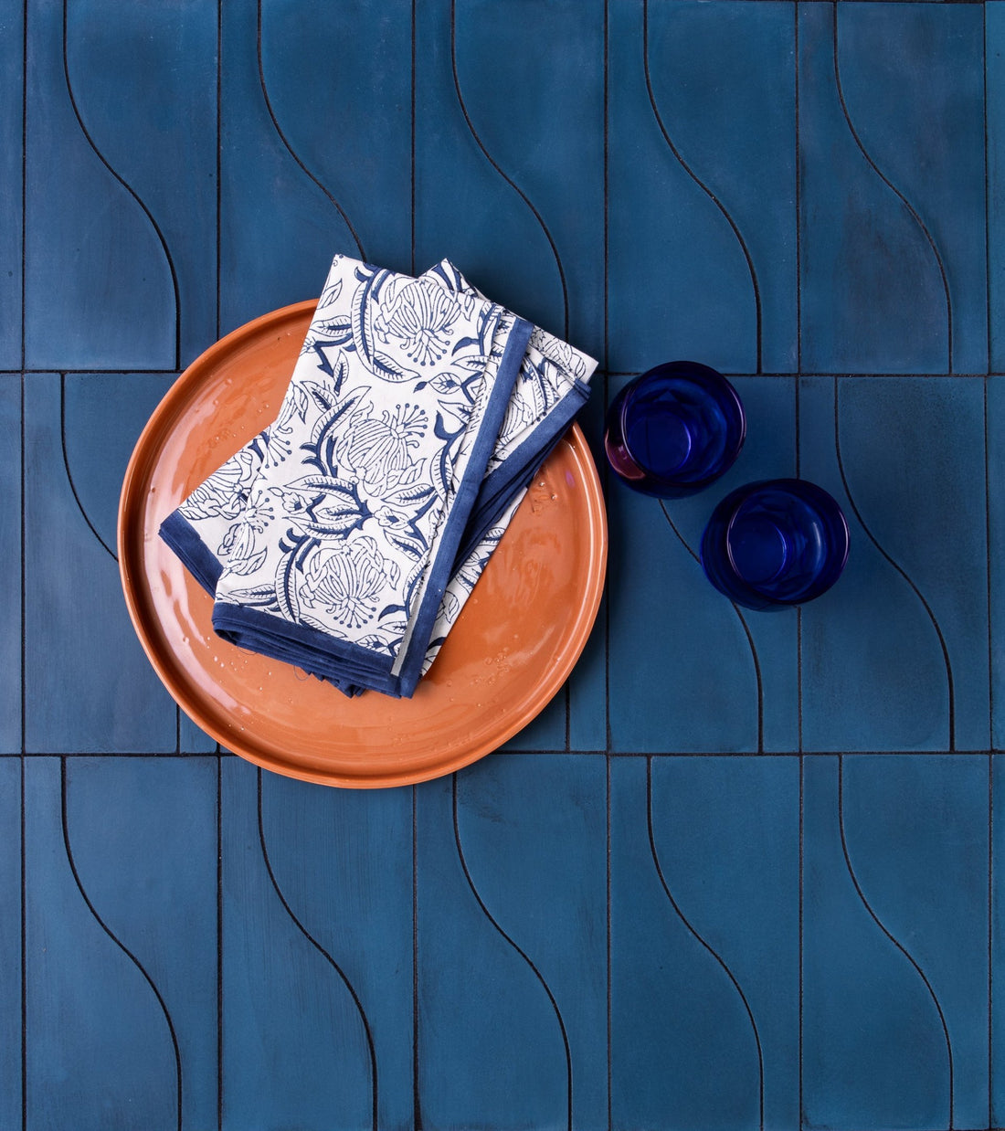 A Nouveau Midnight terracotta plate holds folded white napkins with blue floral patterns, placed on a vendor-unknown Cement tile kitchen backsplash. Nearby are two small dark blue glass cups.