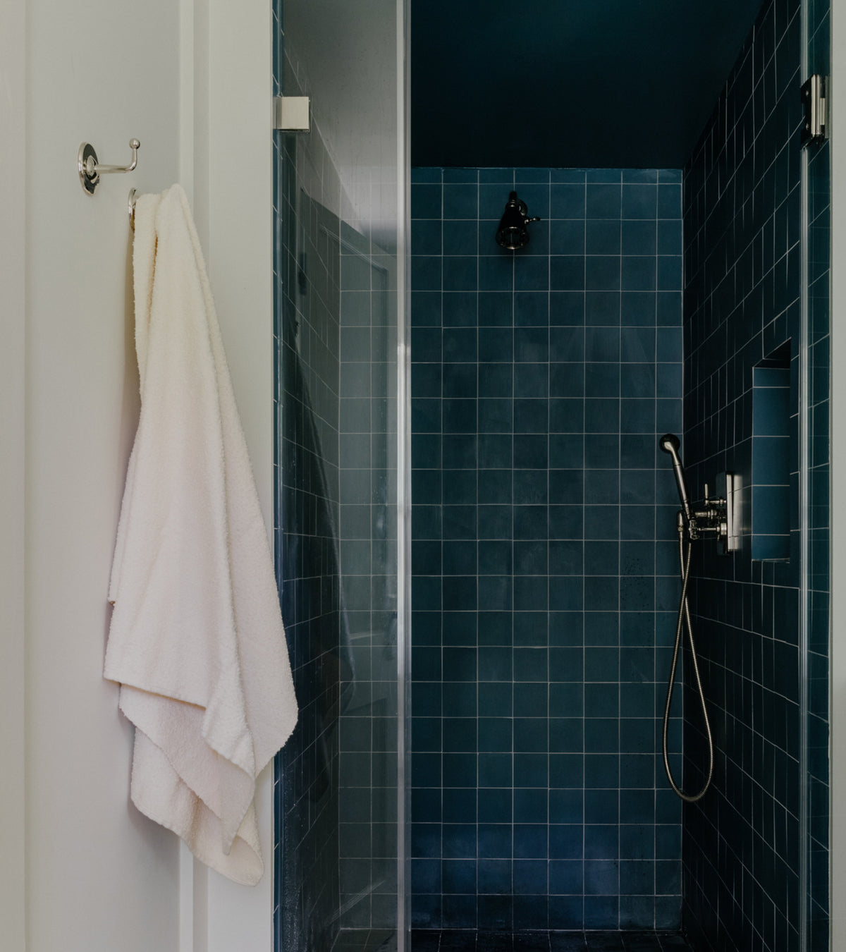 A walk-in shower with Midnight 4x4 cement tile walls, a handheld showerhead, and a white towel hanging on a hook outside the glass door.