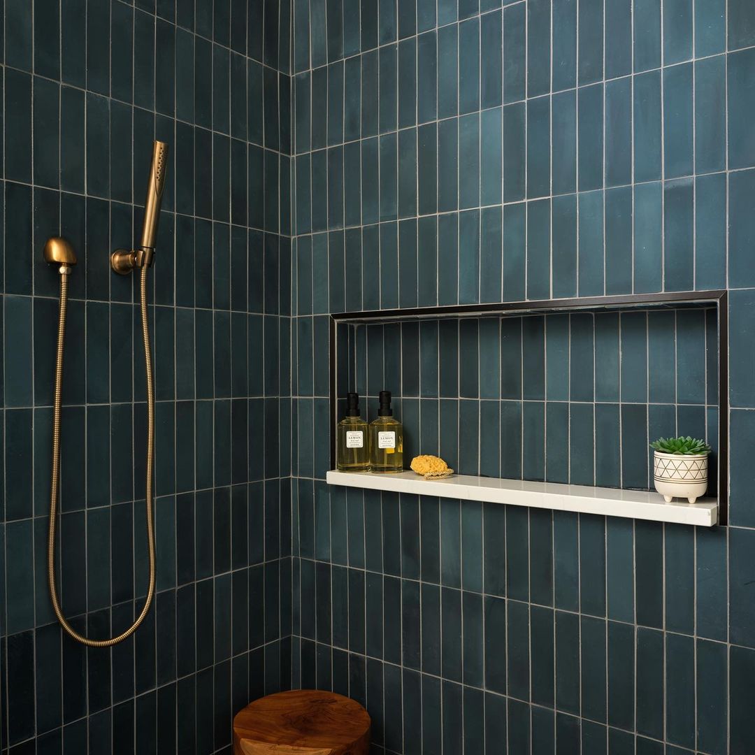 A modern shower features vendor-unknown’s Midnight 2x8 dark blue subway tile, a brass showerhead, wooden stool, and a recessed shelf with bottles, a sponge, and a small potted plant.