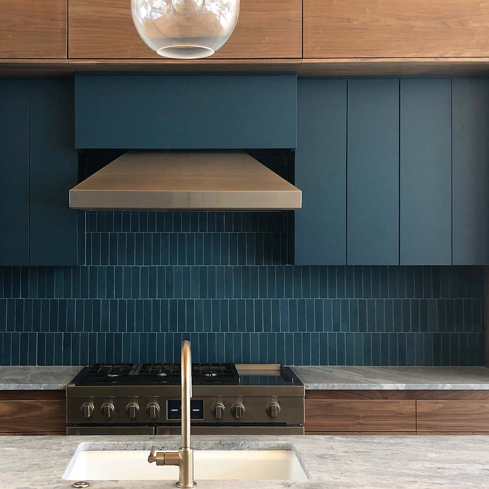 Modern kitchen with vendor-unknown Midnight 2x8 handmade teal backsplash tile, stainless steel gas stove, wood cabinets with matte blue doors, large range hood, marble countertop, gold faucet over white sink, and glass pendant light.