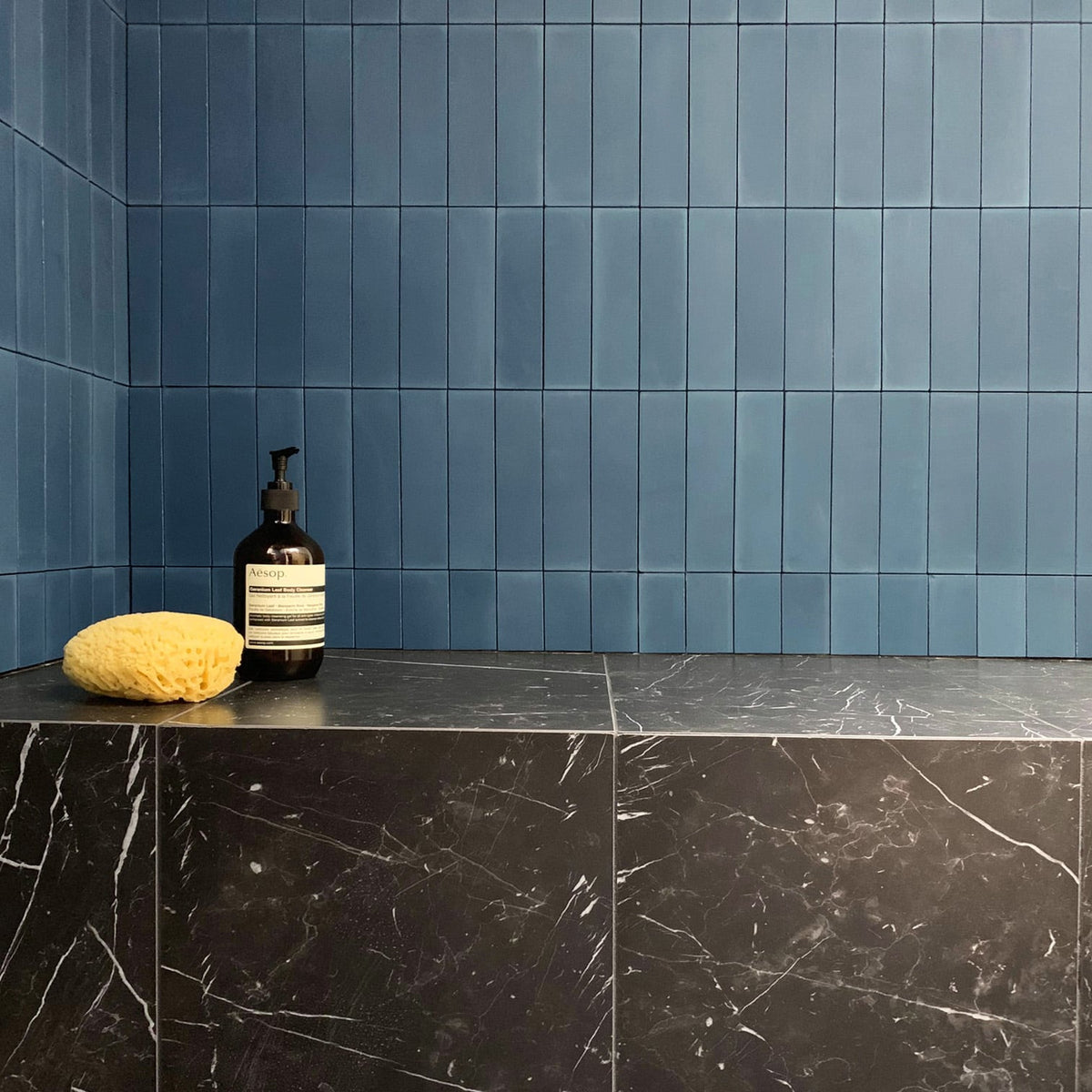 A yellow sponge and a black pump bottle rest on a dark marble ledge below a wall of vendor-unknown Midnight 2x8 blue tile in a modern bathroom.