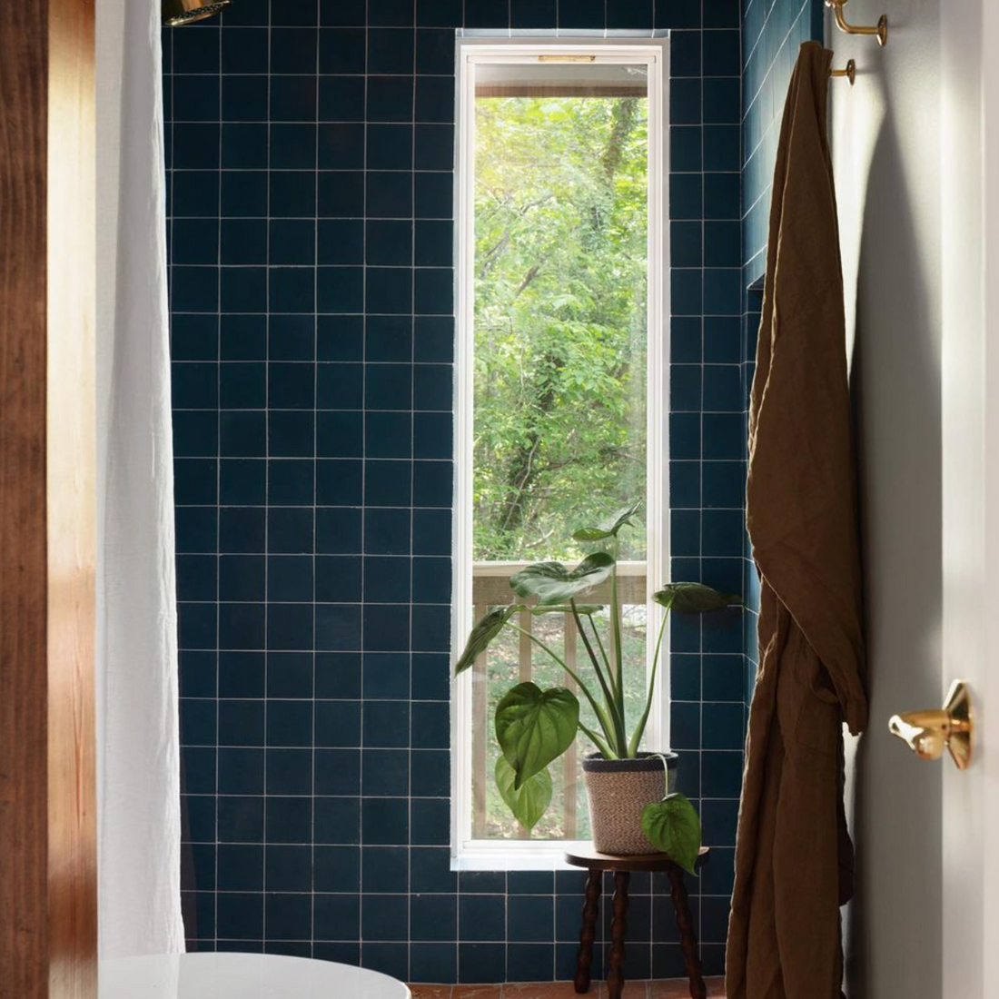 A modern bathroom showcases vendor-unknown’s Midnight 4x4 tiles on the floor, with a tall narrow window for natural light, a leafy potted plant on a stool, and a brown towel hanging from a hook.
