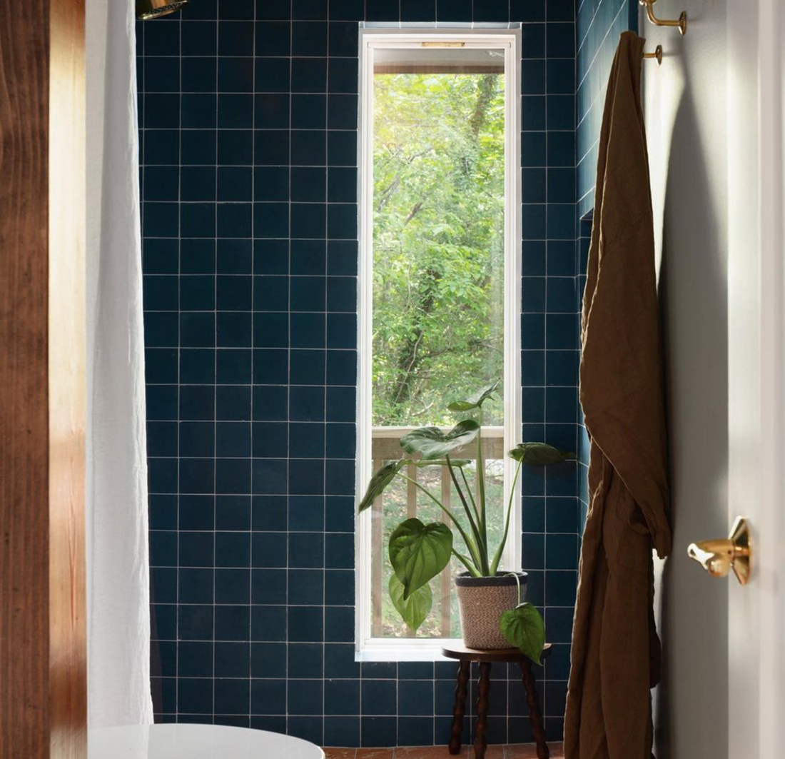 A modern bathroom showcases vendor-unknown’s Midnight 4x4 tiles on the floor, with a tall narrow window for natural light, a leafy potted plant on a stool, and a brown towel hanging from a hook.
