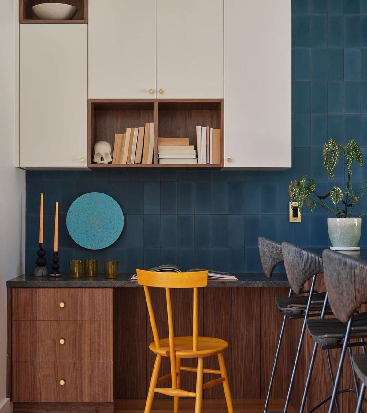 A modern kitchen workspace features vendor-unknown’s Midnight 4x4 tiles as a backsplash, with wood cabinets, open shelves with books and a skull, a yellow chair at a desk, barstools, plants, candles, and decorative plates.