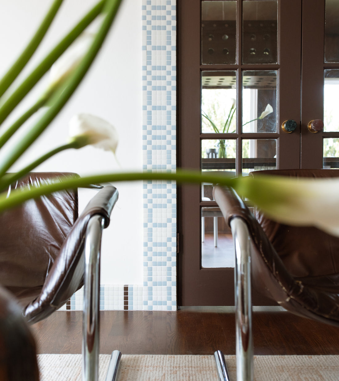 Two brown leather chairs flank a vertical strip of Vaso Milk + Tranquil Blue grid-patterned tiles next to a brown glass-paned door.
