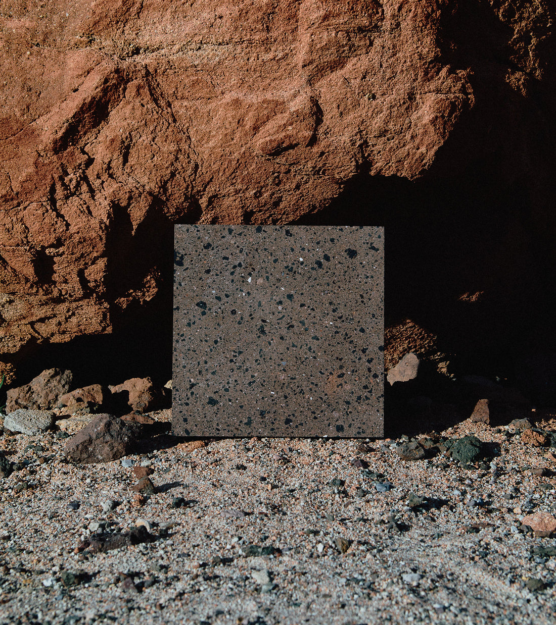 The Moab 12x12 by Zia Tile is a square, dark speckled tile standing upright on sand before a rugged reddish-brown rock wall, illuminated by sunlight.