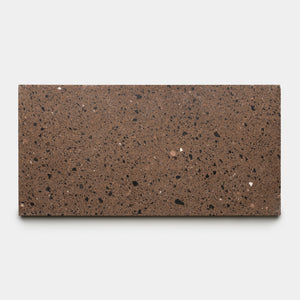 The Moab 12x24 by Zia Tile is a rectangular slab of chocolate brown terrazzo tile featuring small black, dark brown, and white speckles, shown against a plain light gray background.