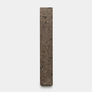 The Zia Tile Moab 2x12 features a tall, rectangular block of speckled brown and black stone, standing upright against a plain light gray background.