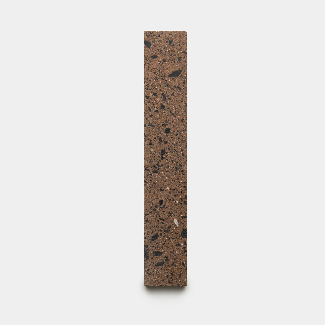 The Moab 2x12 by Zia Tile is a vertically standing rectangular terrazzo block featuring a brown base with scattered black and gray stone fragments, displayed against a plain white background.