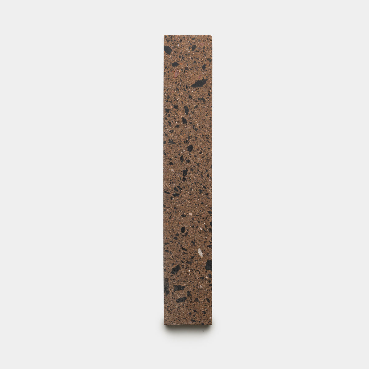 The Moab 2x12 by Zia Tile is a vertically standing rectangular terrazzo block featuring a brown base with scattered black and gray stone fragments, displayed against a plain white background.