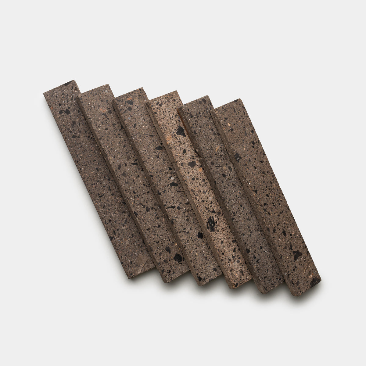 Six Moab 2x12 tiles by Zia Tile, featuring a rectangular, stone-like design with a speckled, porous texture, are arranged diagonally in a staggered row on a plain white background.