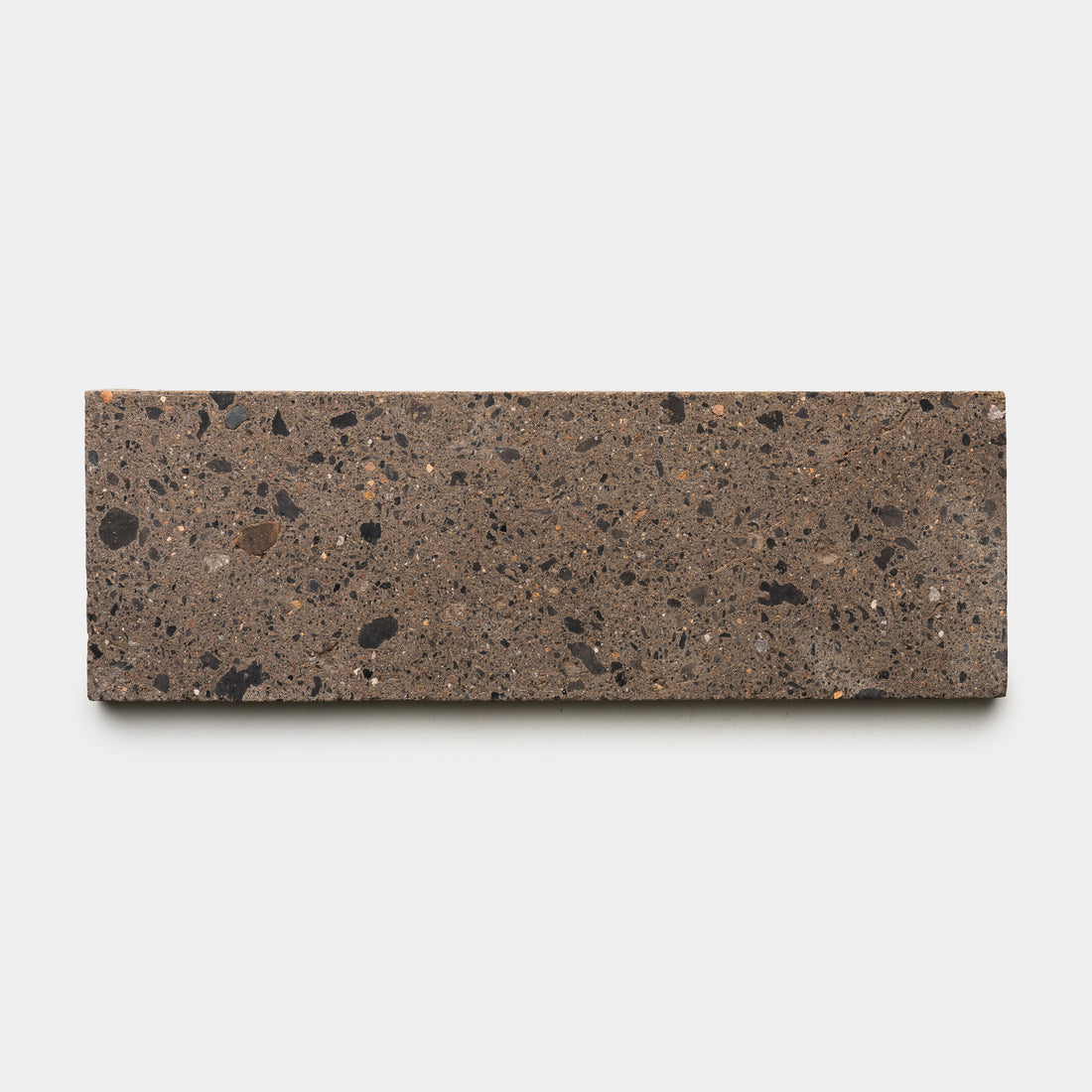 The Moab 4x12 by Zia Tile is a rectangular brown terrazzo ceramic tile with a rough texture and scattered black and dark brown specks, shown against a plain white background—a stylish choice to enhance any room’s character.