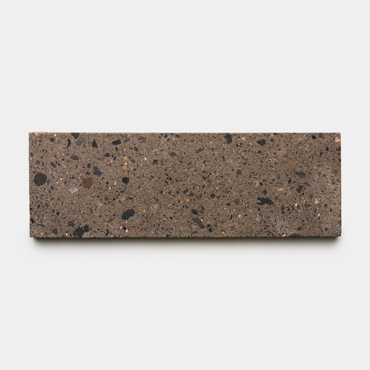 The Moab 4x12 by Zia Tile is a rectangular brown terrazzo ceramic tile with a rough texture and scattered black and dark brown specks, shown against a plain white background—a stylish choice to enhance any room’s character.