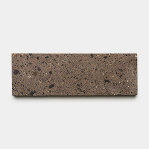 The Moab 4x12 by Zia Tile is a rectangular brown terrazzo ceramic tile with a rough texture and scattered black and dark brown specks, shown against a plain white background—a stylish choice to enhance any room’s character.