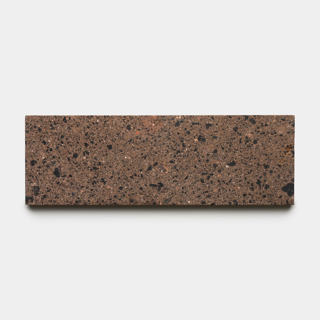 A rectangular brown handmade terrazzo tile featuring black and reddish aggregate pieces, the Moab 4x12 by Zia Tile is displayed against a plain white background.