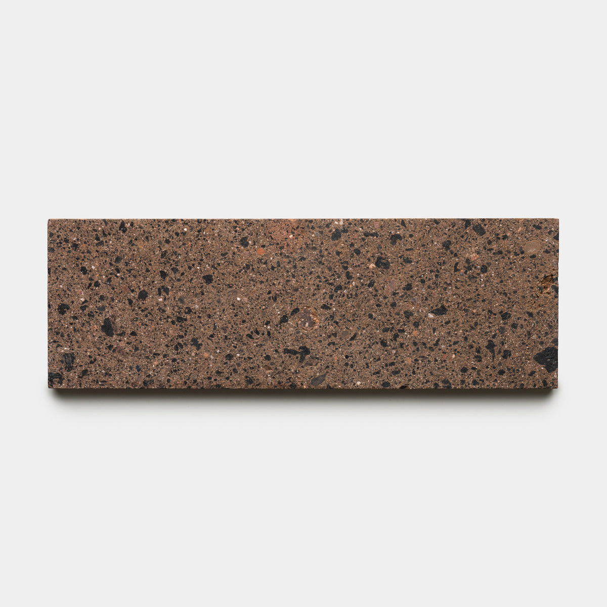 A rectangular brown handmade terrazzo tile featuring black and reddish aggregate pieces, the Moab 4x12 by Zia Tile is displayed against a plain white background.