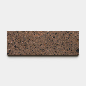 A rectangular brown handmade terrazzo tile featuring black and reddish aggregate pieces, the Moab 4x12 by Zia Tile is displayed against a plain white background.