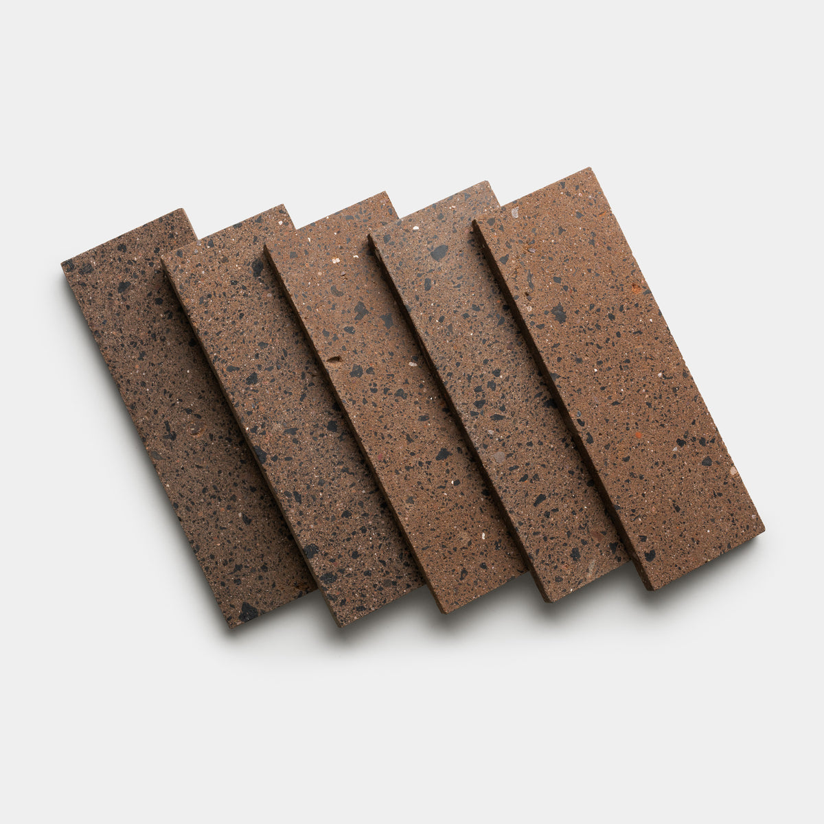 Five rectangular Moab 4x12 brown terrazzo tiles from Zia Tile, featuring black and gray speckles, are arranged in a slightly fanned pattern on a white background, highlighting the craftsmanship of these handmade tiles.