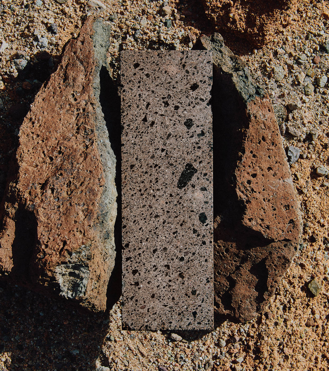 The Moab 4x12 by Zia Tile, a rectangular slab with a handmade look, stands vertically between two rough, reddish-brown rocks on sandy ground, emphasizing an appealing contrast in texture and color.