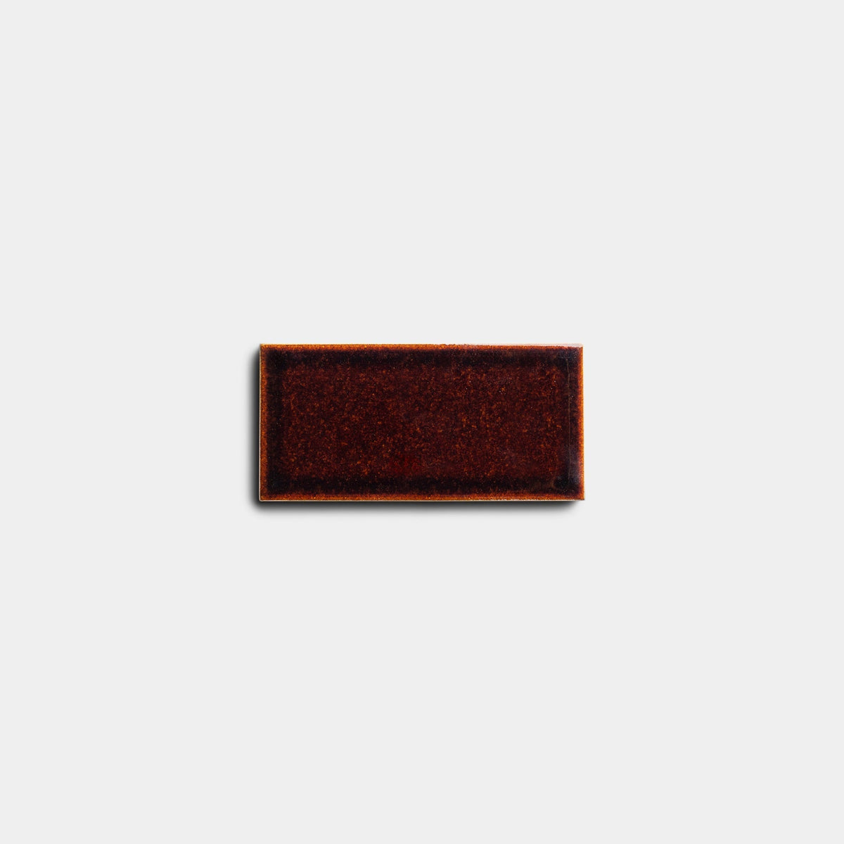 A single Molasses 2x4 ceramic tile sample by Zia Tile, featuring a glossy, dark reddish-brown finish with lighter edges, is displayed against a plain light gray background.