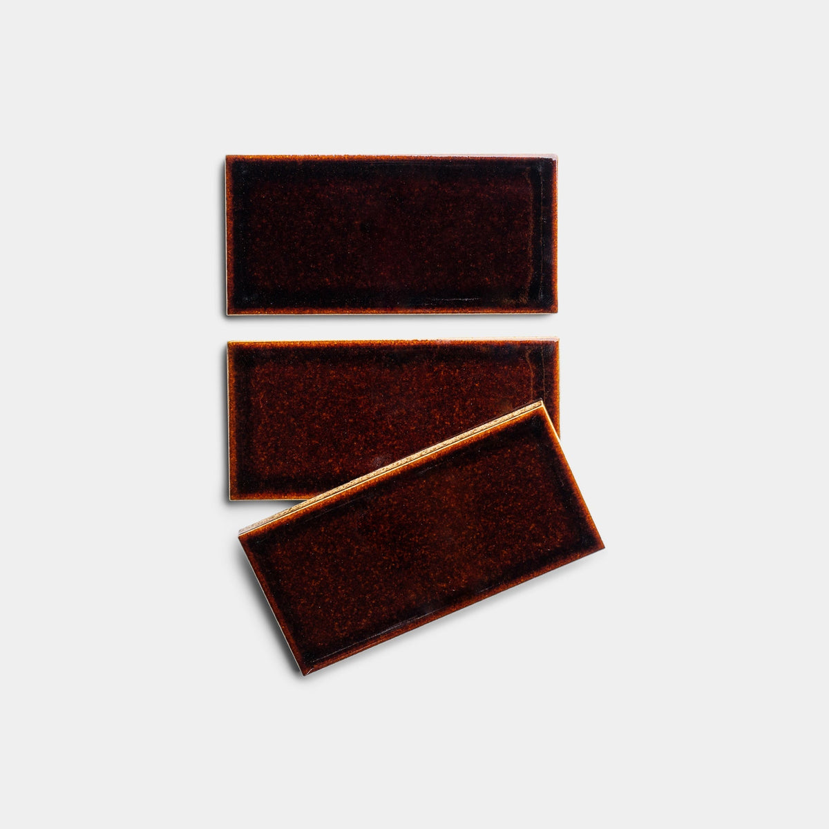 Three Molasses 2x4, Sample tiles by Zia Tile, with a rectangular glossy dark brown finish, are set on a light gray background—two tiles parallel and one slightly tilted to overlap the others’ bottom edges.