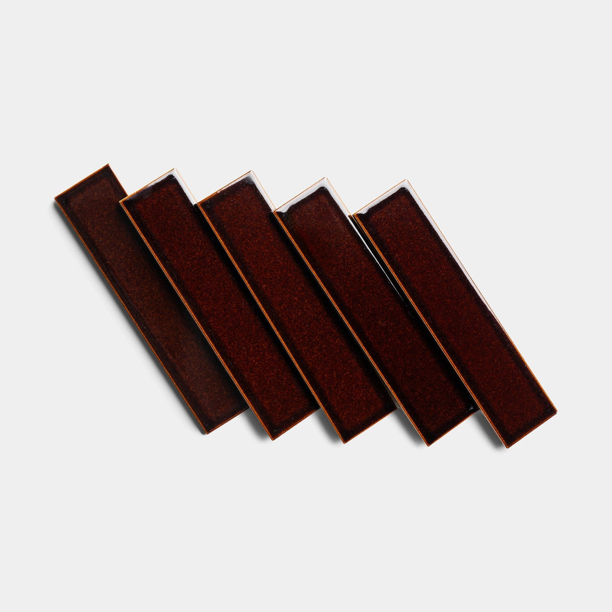 Five rectangular Molasses 2x8 ceramic samples from Zia Tile, dark brown in color, are arranged diagonally in a staggered, overlapping pattern on a plain white background.