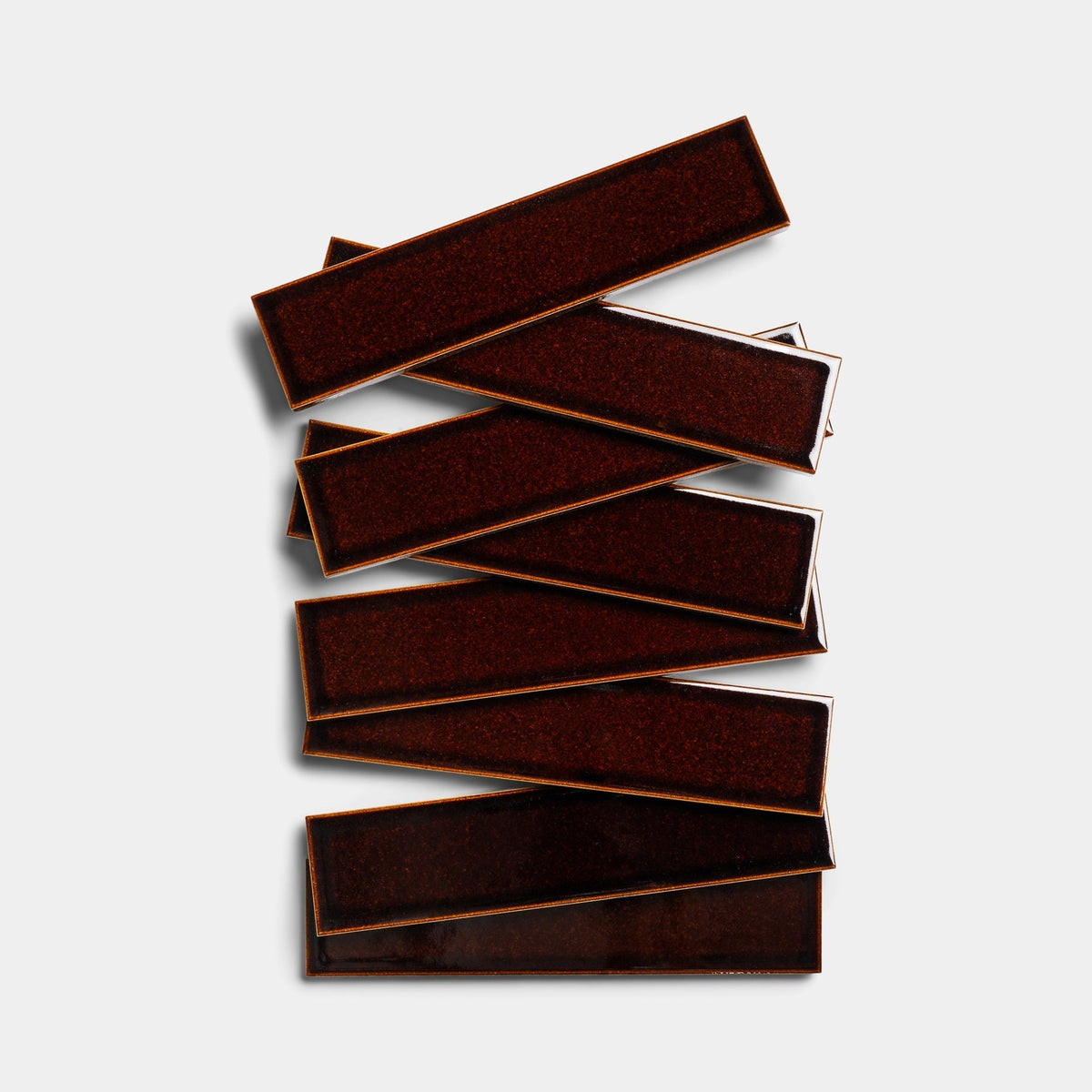 Several Molasses 2x8 sample tiles from Zia Tile, long and dark brown, are stacked in a staggered pattern against a plain white background.