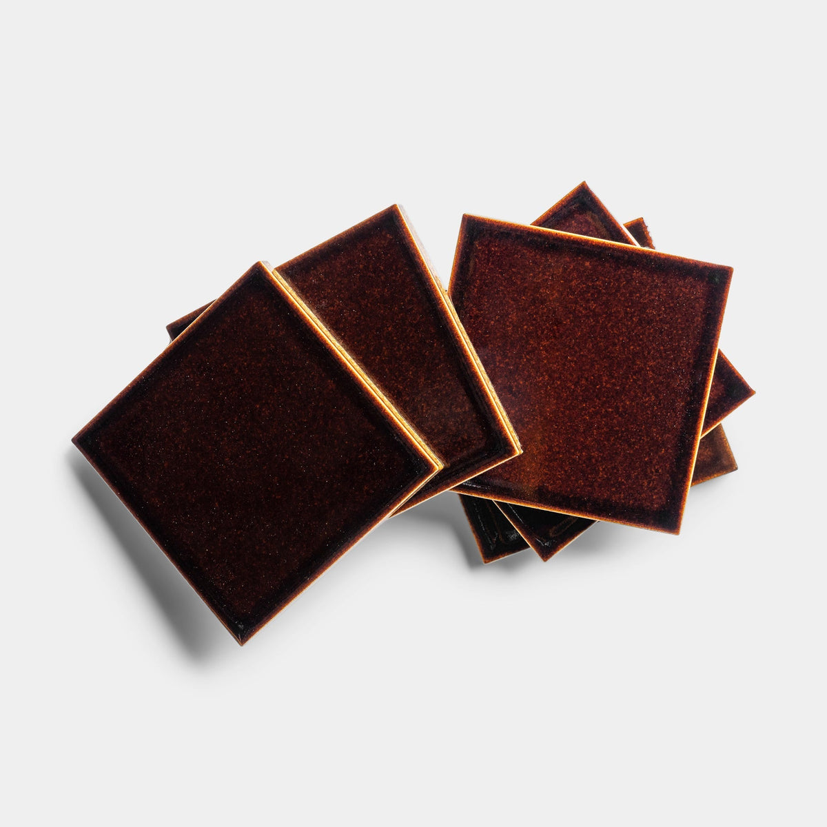 Five Molasses 4x4 ceramic square coasters by Zia Tile, in dark brown with gold edges, are stacked in a slightly scattered arrangement on a white background, casting soft shadows.