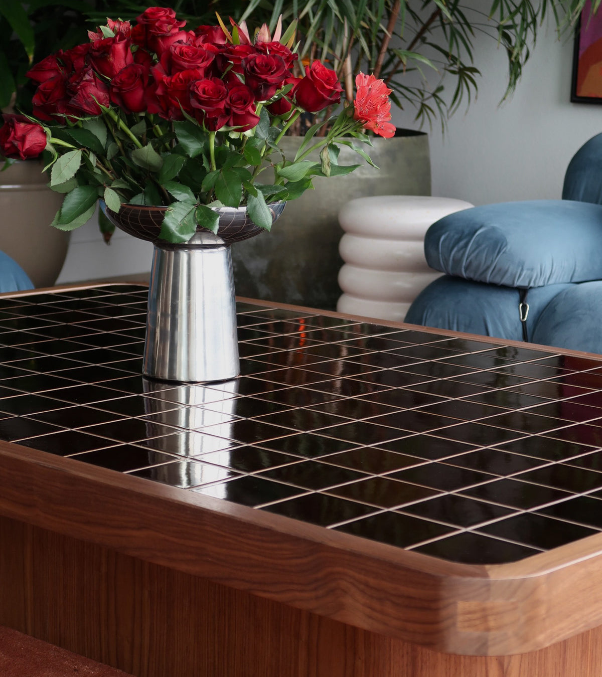 A vase of red roses sits on a Molasses 4x4 glossy black tile coffee table by Zia Tile, featuring a wooden frame, with a plush blue chair and contemporary white stool in the background.