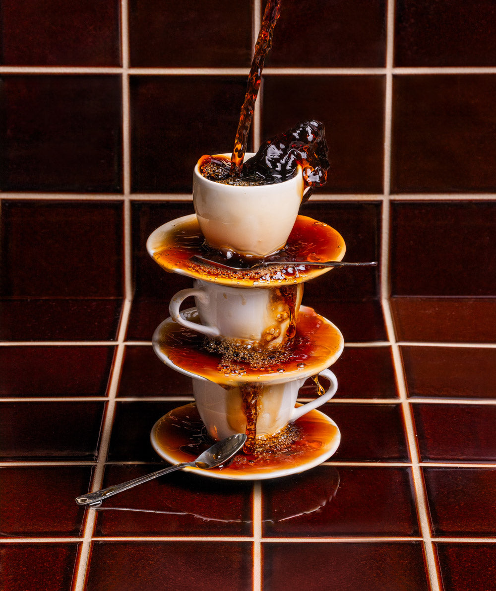 Three stacked white cups and saucers overflow with coffee being poured, spilling liquid down the stack onto a brown tiled surface. A teaspoon rests at the base.
