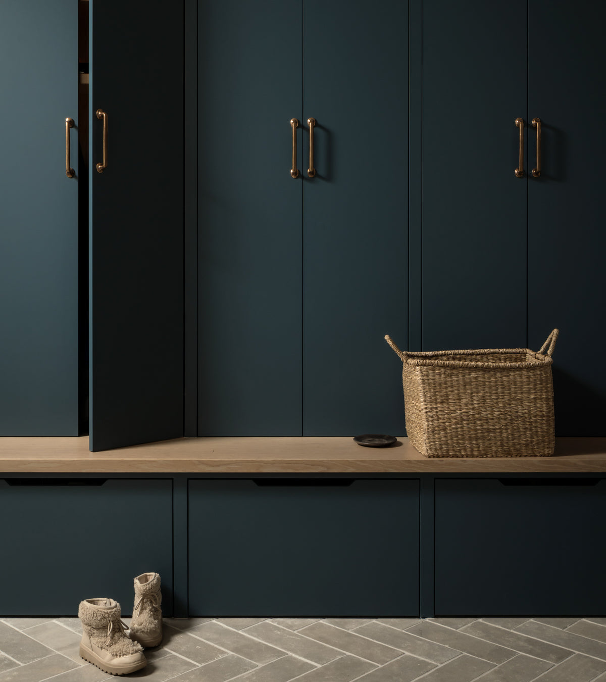 A mudroom with dark blue cabinets, brass handles, a wooden bench, woven basket, and boots on Zia Tile Monument 4x12 + Honed stone tiles in a herringbone pattern. One cabinet door is slightly open.