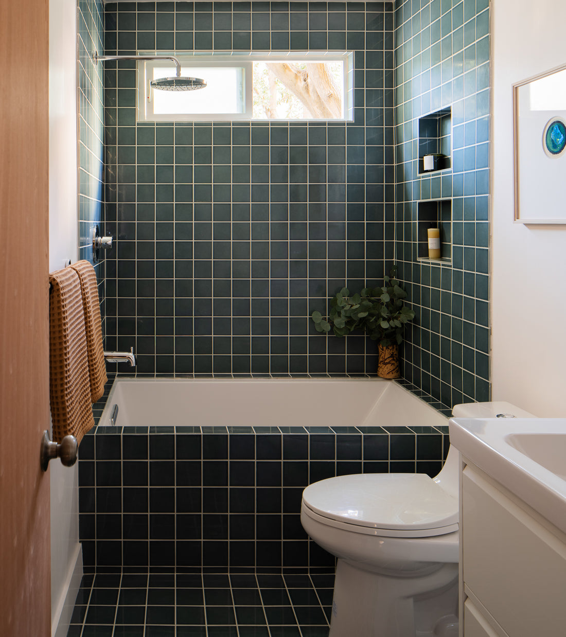A modern bathroom showcases Zia Tile’s Nagano 4x4 dark green ceramic tiles on the walls, tub, and floor. A white toilet, vanity, towel rack, built-in shelves, and a window above the tub complete this Nagano-inspired space.