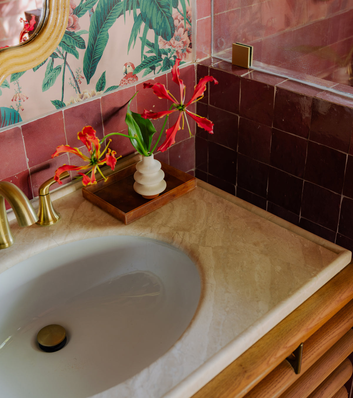 A bathroom vanity featuring deep red Nana’s Lipstick 4x4 Moroccan zellige wall tiles, a light stone countertop with an inset sink, and a small vase with red lily-like flowers.